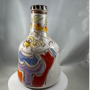 Puede incluir: Un jarrón de cerámica pintado a mano con un diseño único. El jarrón presenta una base blanca con figuras abstractas coloridas en rojo, naranja, morado y azul. El cuello del jarrón tiene un borde negro y marrón, con detalles en amarillo y verde.