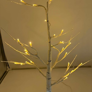 Könnte beinhalten: Ein dekorativer LED-Birkenbaum mit warmweißen Lichtern. Der Baum hat einen weißen Stamm mit schwarzen Markierungen und dünnen Ästen. Die Lichter sind gleichmäßig über die Äste verteilt und erzeugen ein sanftes Leuchten. Der Baum ist eine festliche Dekoration.
