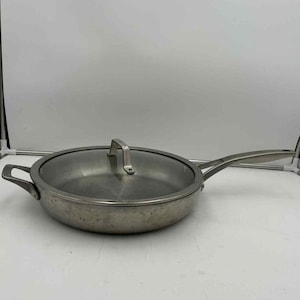 Puede incluir: Sartén de acero inoxidable con tapa de cristal. La sartén tiene un mango largo y otro más pequeño en el lado opuesto. La tapa tiene un asa de acero inoxidable. La sartén tiene un diámetro de unos 30 cm.