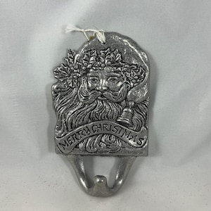 Puede incluir: Un porta calcetines de Navidad de metal plateado con una escultura en relieve de la cara de Papá Noel con una corona de acebo y bayas. El porta calcetines tiene un gancho en la parte inferior y dice "Feliz Navidad" en una pancarta.