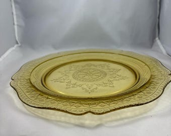 May include: A yellow glass plate with a scalloped edge and a delicate floral pattern. The plate has a raised center with a circular design.