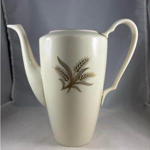 May include: Cream-colored ceramic coffee pot with a gold wheat stalk design. The pot has a long, curved spout and a handle with a unique, open design. The wheat design is centered on the pot's body.