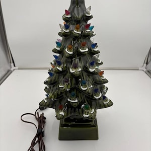 Może przedstawiać: Ceramiczne drzewko bożonarodzeniowe z błyszczącym zielonym wykończeniem. Drzewko zdobią kolorowe, plastikowe żarówki i białe akcenty. Ma kwadratową podstawę i brązowy przewód zasilający.
