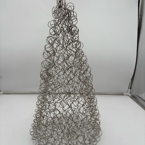 May include: A silver-colored, cone-shaped sculpture made of intertwined wire. The sculpture has a rounded top and a textured surface created by the overlapping wire loops. The design resembles a stylized Christmas tree.