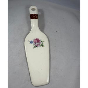 Pode incluir: Uma espátula de bolo de cerâmica branca com um design floral. A alça apresenta um padrão marrom decorativo e a lâmina é adornada com rosas rosa, roxas e azuis e folhas verdes.