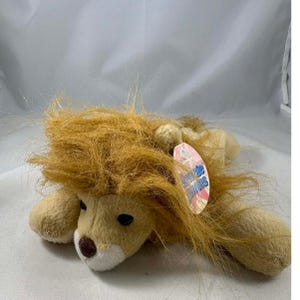 Peut inclure: Un jouet en peluche en forme de lion avec un corps beige, un museau blanc et un nez marron. Le lion a une crinière hirsute brun clair et des yeux noirs. Une étiquette est attachée au jouet.