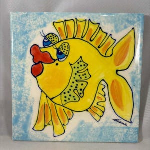 May include: A square ceramic tile featuring a whimsical yellow fish illustration. The fish has large blue eyes, a red mouth, and orange accents. The background is a light blue with white accents. The artist's signature, "diane", is in the lower right corner.