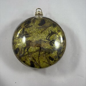 May include: A round, clear glass Christmas ornament with a gold top. The ornament contains a glittery, green background with a silhouette of a deer.