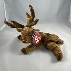 Puede incluir: Un reno de peluche marrón con la nariz roja y grandes cuernos. El reno está tumbado con la cabeza girada hacia un lado. Tiene una etiqueta Ty unida a su pierna.