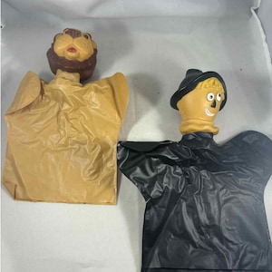 Könnte beinhalten: Zwei Vintage-Handpuppen: ein Löwe mit brauner Mähne und eine Vogelscheuche mit schwarzem Hut. Die Löwenpuppe hat einen beigefarbenen Körper, während die Vogelscheuche einen schwarzen Körper hat. Beide Puppen haben bemalte Gesichter und sind aus weichem Material.