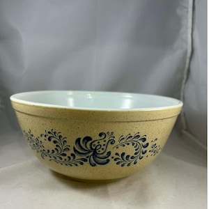 May include: A tan mixing bowl with a speckled finish and a white interior. The bowl features a blue floral design around the exterior. This vintage bowl is suitable for kitchen use.