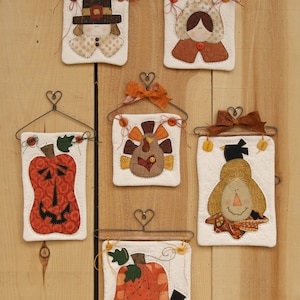 May include: Six handmade felt wall hangings with a Thanksgiving theme. Each hanging features a different design, including a pilgrim, a turkey, a pumpkin, and a scarecrow. The hangings are all white with brown, orange, and red accents.