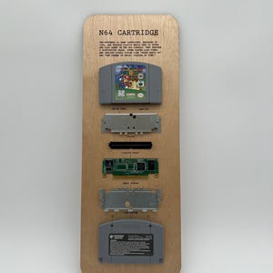 May include: A wooden display board featuring an N64 cartridge and its disassembled components. The gray cartridge displays a colorful game label. The board includes text describing the cartridge's history and features.
