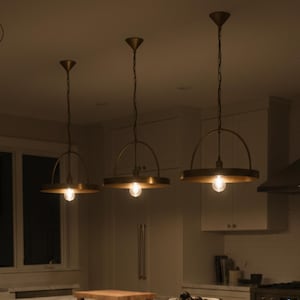 Puede incluir: Tres lámparas colgantes con pantallas de metal color latón y bombillas expuestas cuelgan sobre una isla de cocina. Las luces están suspendidas del techo por cadenas. La cocina cuenta con gabinetes blancos, una encimera de mármol y una campana extractora de acero inoxidable.