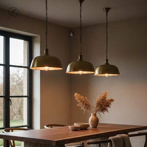May include: Three brass pendant lights hang above a wooden dining table. The lights have a dome shape and are suspended by chains. A vase with dried plants and two bowls are on the table. The room has a neutral colour palette with a large window.