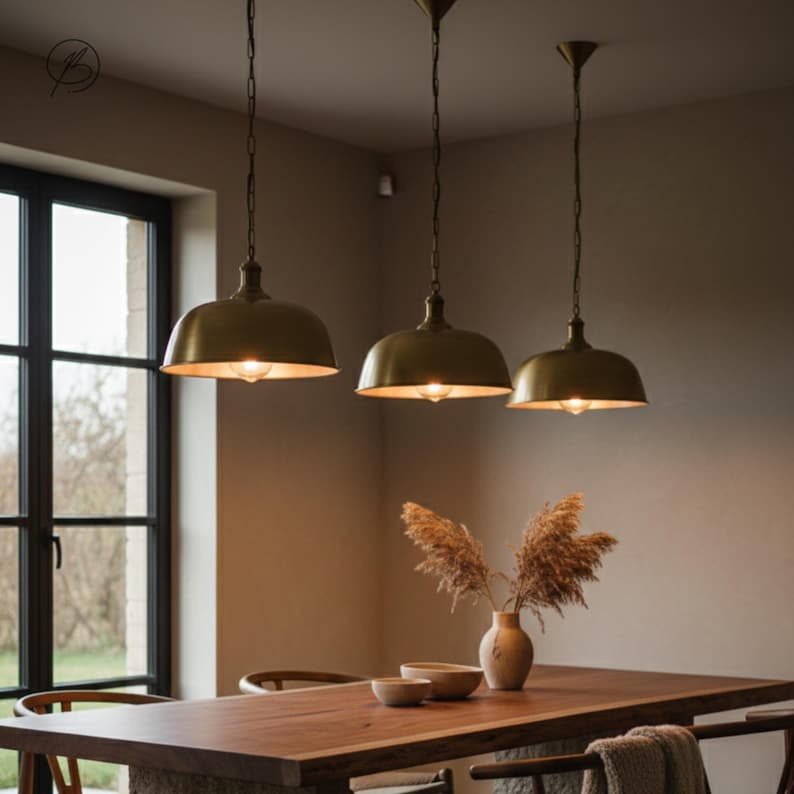 May include: Three brass pendant lights hang above a wooden dining table. The lights have a dome shape and are suspended by chains. A vase with dried plants and two bowls are on the table. The room has a neutral colour palette with a large window.
