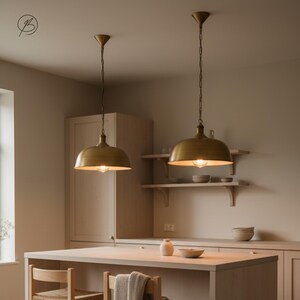 May include: Two brass-coloured pendant lights suspended from the ceiling. Each light features a rounded shade and a visible lightbulb. The lights hang above a light-coloured wooden kitchen island. The kitchen has light-coloured cabinets and shelves.