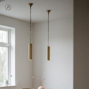 May include: Two brass cylindrical pendant lights suspended from the ceiling by chains. Each light has a conical top. The lights are in a room with a window and a vase with dried flowers. The room has a neutral colour scheme.