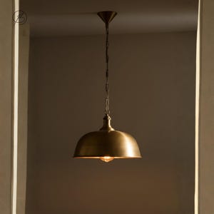 May include: A brass pendant light with a domed shade, suspended by a chain from a conical ceiling fitting. The light is on, emitting a warm glow. The design is reminiscent of industrial lighting.