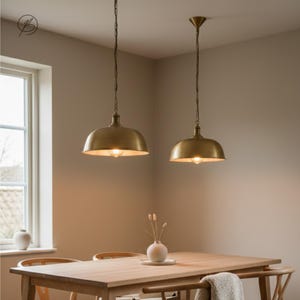 May include: Two brass-toned pendant lights suspended from the ceiling above a wooden dining table. The lights have a domed shape and are hanging by chains. The room has a neutral colour scheme, with a window and a vase of flowers on the table.