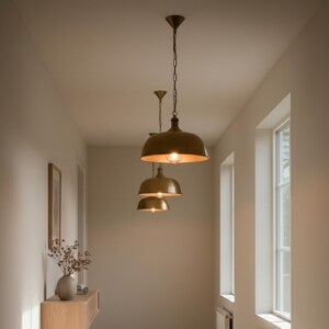 May include: Three brass pendant lights hanging in a hallway. The lights have a dome shape and are suspended from chains. The hallway has neutral-coloured walls and a wooden console table with a vase of dried flowers.