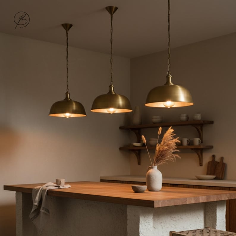 May include: Three brass pendant lights suspended over a wooden kitchen island. Each light has a dome-shaped shade and a single exposed bulb. The kitchen features a neutral colour palette with wooden shelves and a vase of dried flowers.