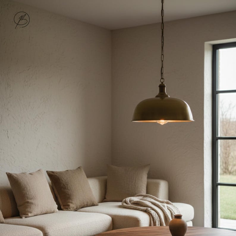 May include: A bronze pendant light suspended from a chain in a living room. The light has a rounded, dome-shaped shade and a single exposed bulb. The room features a neutral colour palette with a cream sofa, throw cushions, and a wooden coffee table.