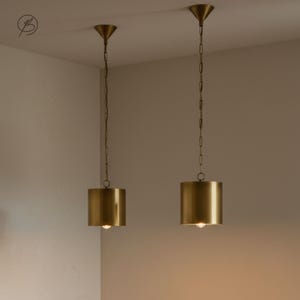 Może przedstawiać: Dwie cylindryczne lampy wiszące z mosiężnym wykończeniem wiszą pod sufitem. Każda lampa jest zawieszona na łańcuchu i ma stożkowy daszek. Światła są zapalone, emitując ciepłą poświatę.