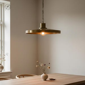 May include: A brass-colored pendant light fixture with a circular shade hangs from a chain. The light bulb is visible, emitting a warm glow. The fixture is suspended above a wooden table, with a vase of flowers in the foreground.