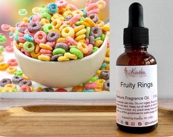 May include: A bowl of colorful cereal with a spoon, next to a brown glass bottle of "Fruity Rings" premium fragrance oil. The bottle has a black dropper and a white label with text and a floral design. The cereal is in a white bowl on a wooden tray.