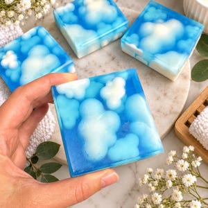 May include: Four square bars of soap featuring a translucent blue base with embedded opaque white cloud shapes. The soap has a smooth, glossy finish and a distinct cubic form, showcasing a clear sky pattern with fluffy white clouds throughout.