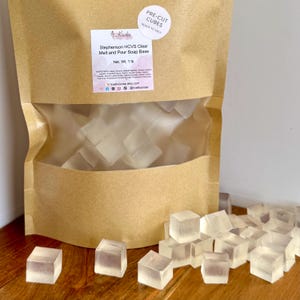 May include: A brown paper bag filled with clear, pre-cut soap cubes. The bag has a clear window showing the cubes inside. A label reads "Stephenson HCVS Clear Melt and Pour Soap Base" and "Net. Wt. 1 lb". The cubes are scattered on a wooden surface.