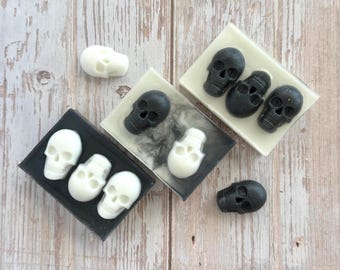 May include: A collection of skull-shaped soaps in black and white, arranged on small square soap dishes. The soaps are displayed on a light wooden surface, creating a spooky and decorative theme.