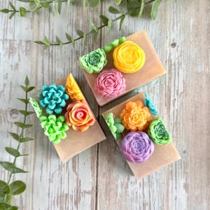 May include: Three rectangular bars of soap, each adorned with colorful, sculpted floral designs. The soaps feature a variety of flower shapes in shades of yellow, pink, orange, blue, and purple, set against a light brown background. The soaps are arranged on a wooden surface.
