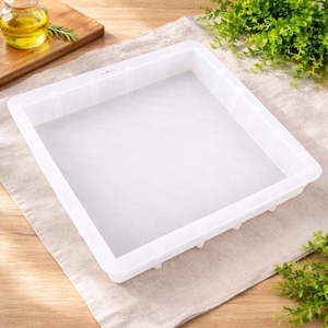 May include: A white rectangular soap mold with a flat bottom and slightly raised sides. The mold is empty and sits on a beige linen surface. A small bottle of oil and greenery are in the background.