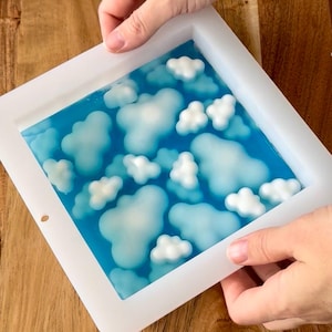 May include: A white rectangular mold filled with blue resin and white cloud-shaped embellishments. The mold is held by hands, showcasing the cloud-filled resin art. The resin art resembles a sky scene.