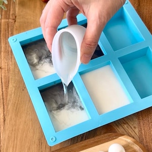 May include: A light blue silicone soap mold with six rectangular cavities. A white cup is pouring a clear liquid into the mold. Some cavities are filled with white and gray soap. The mold is on a wooden surface.