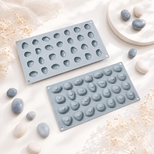 May include: Two rectangular light blue silicone molds featuring multiple pebble-shaped cavities. One mold displays the concave indentations, while the other shows the convex raised shapes. Several smooth, oval-shaped stones in blue and white are scattered.