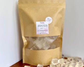 May include: A brown paper bag with a clear window, filled with clear, pre-cut cubes. The bag has a label that reads "Stephenson HCVS Clear Melt and Pour Soap Base." A sticker on the bag says "PRE-CUT CUBES." Cubes are scattered on a wooden surface.