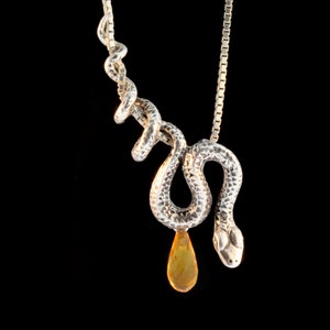 Puede incluir: Un collar con un colgante de serpiente de plata con una piedra preciosa de ámbar en forma de lágrima que cuelga de su cola.