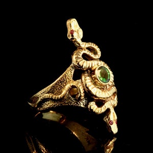 Pode incluir: Um anel de ouro com um design detalhado de duas cobras entrelaçadas. As cobras têm olhos vermelhos e uma gema verde no centro.