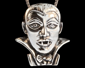 May include: A silver pendant necklace featuring a detailed sculpted face of a vampire with fangs, wearing a ruffled collar.