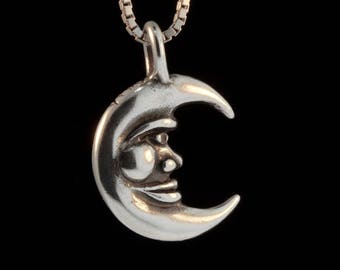 Puede incluir: Un colgante de plata en forma de media luna con una cara. El colgante está colgado de una cadena de plata.