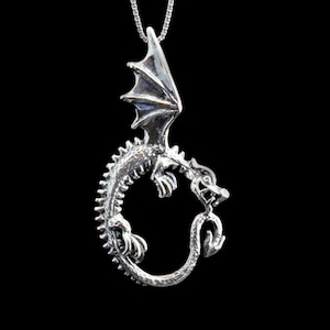 Op de afbeelding: Een zilveren drakenhangerketting met de draak opgerold in een cirkel en zijn vleugels uitgespreid. De draak is gedetailleerd met schubben en klauwen.