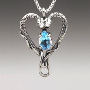 Op de afbeelding: Een zilveren ketting met een hanger met twee slangenhoofden die een hart vormen met een blauwe topaas edelsteen in het midden.