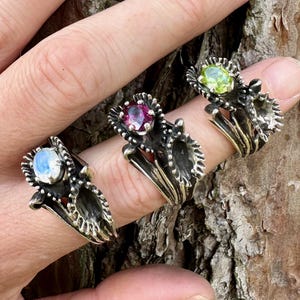 Puede incluir: Cuatro anillos de plata con piedras preciosas de diferentes colores. Los anillos presentan un delicado diseño floral con intrincados detalles. Las piedras preciosas son una piedra de luna azul pálida, un rubí rosa intenso, un peridoto verde claro y un cristal transparente.