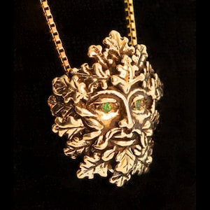 Op de afbeelding: Een gouden ketting met een gedetailleerd Green Man-ontwerp met groene edelsteenogen. De hanger is omgeven door eikenbladeren.