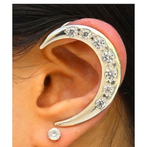 May include: A silver ear cuff with a crescent moon shape and clear rhinestones. The cuff is worn on the upper part of the ear. A small, clear, round stud earring is worn in the lower lobe of the ear.