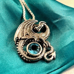 Puede incluir: Un collar con un colgante de plata en forma de dragón y una piedra preciosa azul. El dragón está enroscado alrededor de la piedra, con las alas extendidas. El collar está sobre un fondo de satén azul turquesa.