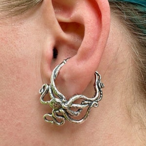 May include: A silver-colored earring featuring an octopus design. The earring is a hoop style, with the octopus's tentacles wrapping around the hoop. The earring is worn in the ear.
